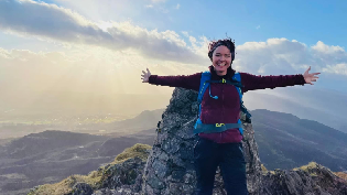 Jo Hedley celebrates reaching the top of a Munro. She smiles with her arms outstretched at the peak with blue skies and the sun shining behind her.