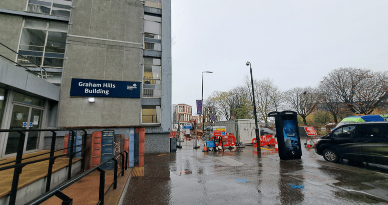 “Graham Hills Building in Glasgow on a rainy street with construction barriers and a black van nearby.”