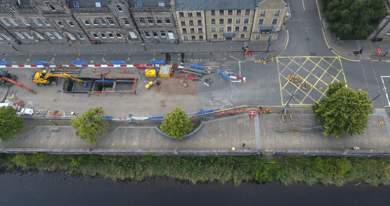An aerial shot of the road surface with excavators next to a dug-up section of the road and pipes underneath. The site is surrounded by fencing and there are buildings on one side, trees and river on the other.