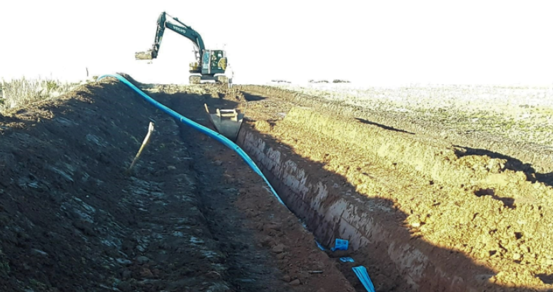 A trench is dug in the ground with a blue pipe running through it. Above ground is a construction vehicle.