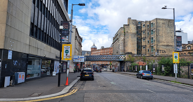 This image shows London Road in Glasgow. There are parked cars on either side of the road and, further down the road, a line of traffic beneath a railway bridge