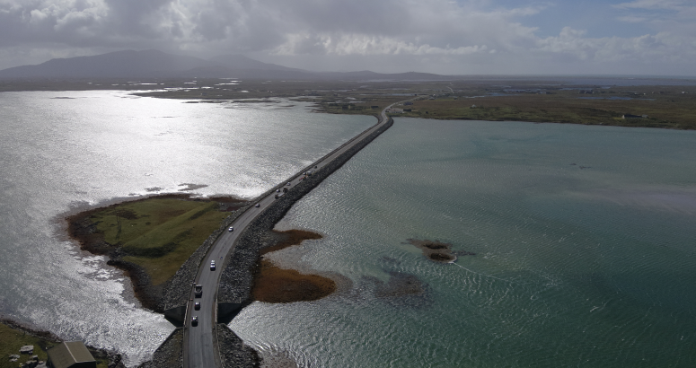 Benbecula Drone Footage Aerial view of a road curving through a coastal landscape with sunlit water and distant mountains.