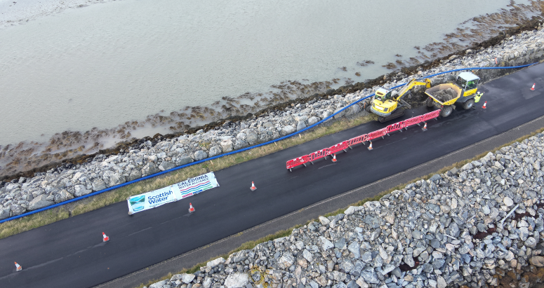 Drone Footage Aerial view of road construction near a water body with vehicles and equipment, featuring a "Caledonia Water Alliance" banner.