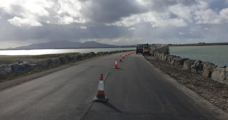 Benbecula Drone Footage Road construction in progress along a coastal road with traffic cones lining one side and machinery ahead under a cloudy sky.