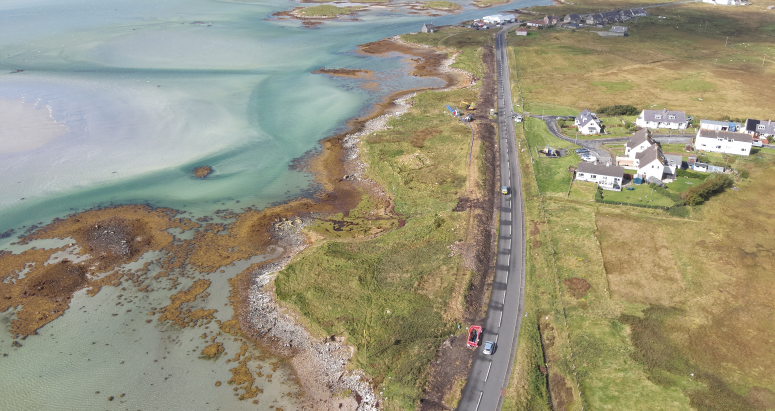 Benbecula Drone Footage Aerial view of a coastal road in Benbecula, Scotland, with adjacent houses and clear turquoise waters alongside rocky shorelines.