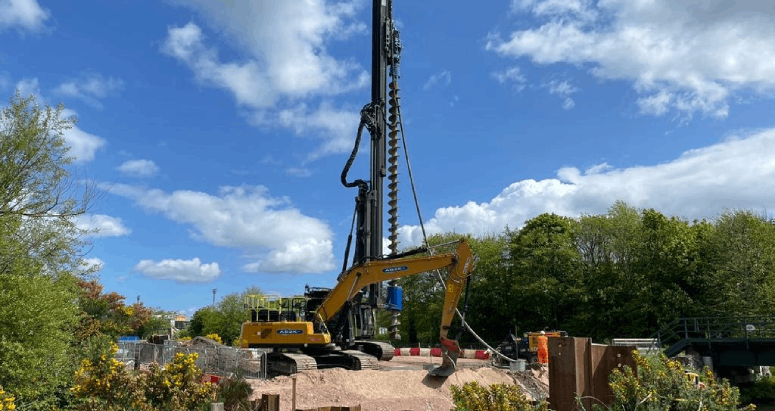 Blue skies and trees/greenery are in the background of this pic. In the centre is heavy machinery with a large drill for piling works. A yellow excavator sits in front of it.
