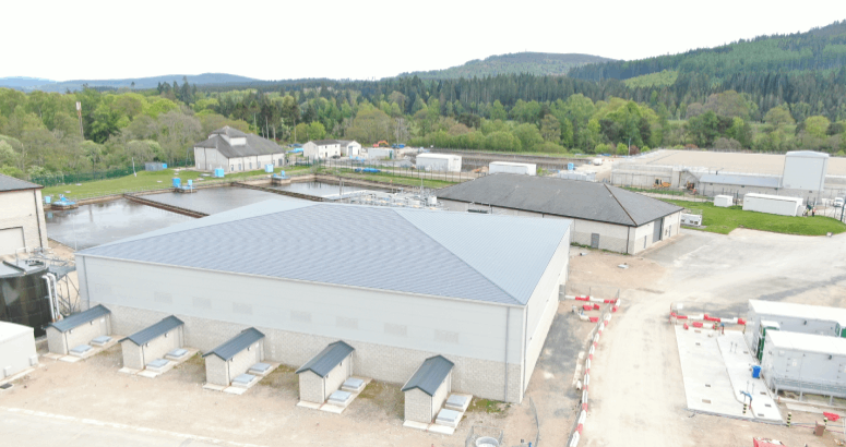 a drone picture showing an industrial works building with construction work happening to the right hand side of the picture. To the left hand side there are water tanks, and in the background greenery and hills.