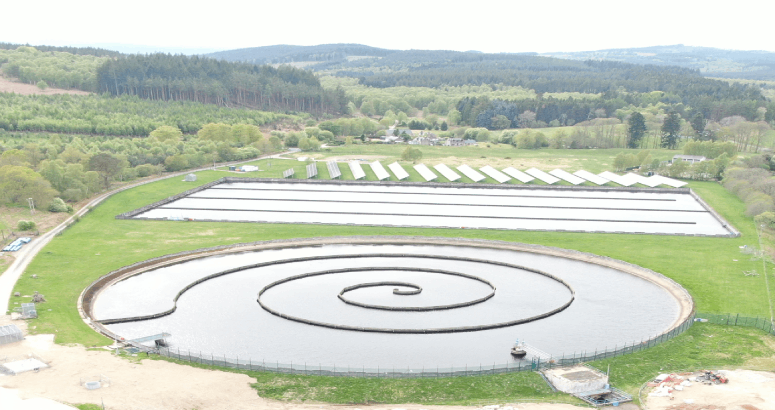 An aerial shot of a spiral water tank in the ground, with grass surrounding it. In the background there are rows of PV solar panels, facing to the left of the picture.