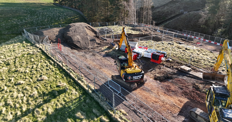 Gladhouse Reservoir Site Compound with George Leslie Excavators at Work Aerial view of a construction site with several excavators, fenced-off areas, and construction materials scattered around, set in a grassy, slightly forested area.