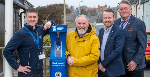 An image taken outdoors, on the left of the blue Scottish Water top-up tap is a Scottish Water employee resting his arm on the top of the tap. On the right of the tap are three community representatives.