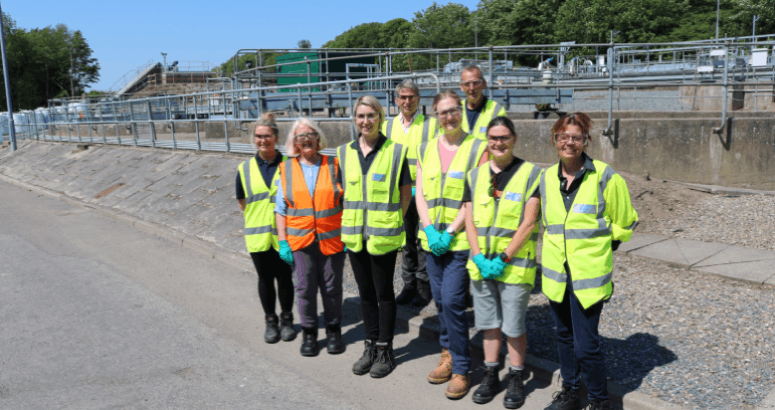 A group of people standing facing the camera wearing hi-vis vests and other PPE. They are standing outside in on a concrete road surface.