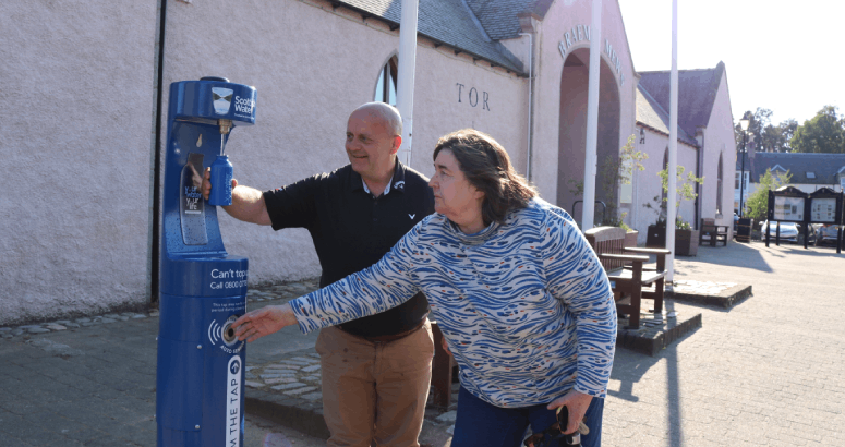 Two people are standing outside a building, holding a blue water bottle underneath a Scottish Water branded water fountain