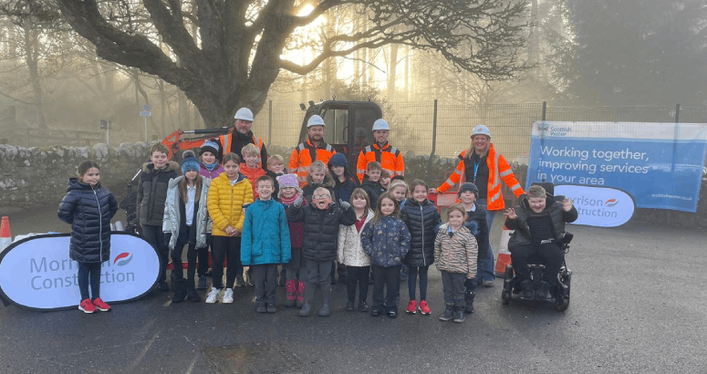 A group of children are standing outside in the school playground, facing the camera. There are four adults wearing orange hi-vis jackets behind them.