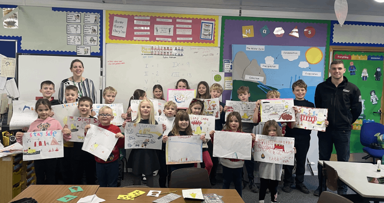 A group of children are standing in a classroom facing the camera. They are holding up posters they have created, and there are two adults standing behind them.