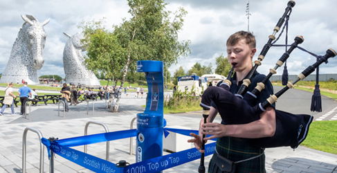 Bagpiper standing next to water fountain with Kelpies sculpture in background