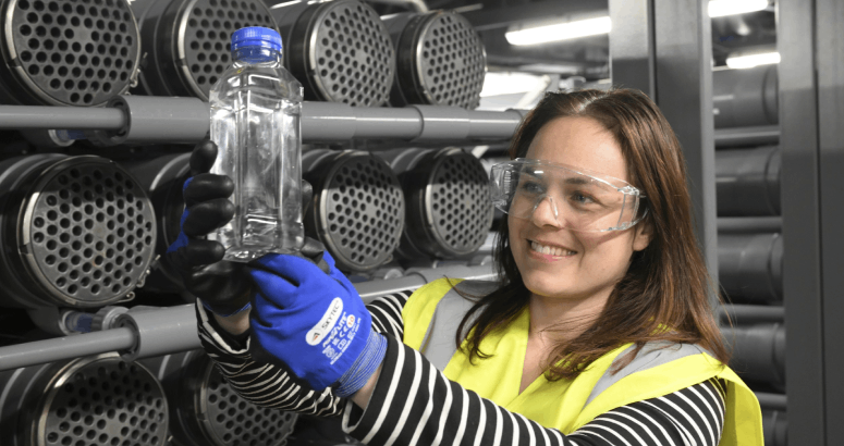 Kate Forbes MSP is standing in frame in an industrial setting, wearing PPE including safety goggles, gloves and hi-viz. She is holding a plastic bottle filled with treated Scottish water.