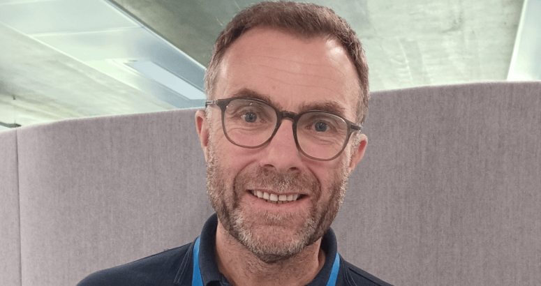 This is a cropped head and shoulders picture of a man called Simon Parsons, who works at Scottish Water. He has stubble and is wearing black-rimmed glasses. He is wearing a dark blue polo short with a lighter blue lanyard round his neck.