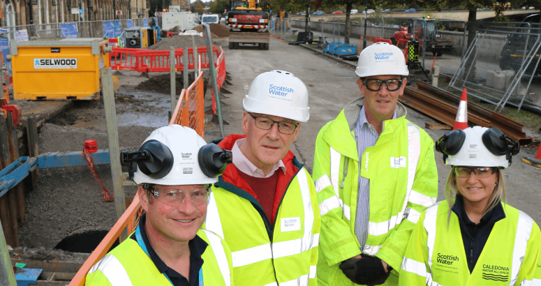 A group of four people wearing PPE including hi-vis, safety goggles, hard hats and gloves, stand in a construction site facing the camera. To the left and in the background are excavations and open pipework.