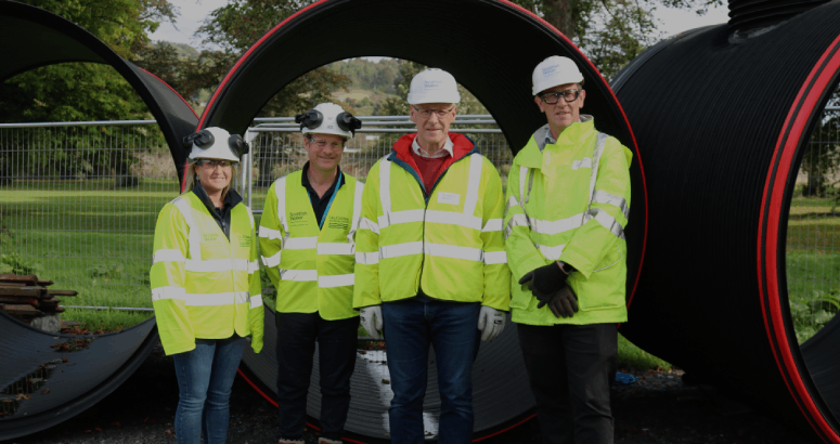 A group of four people wearing PPE including hi-vis, safety goggles, hard hats and gloves, stand outside in front of pipe sections of 2.2 metre diameter. In the background is a grassed area.