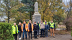 A group of people wearing hi-vis jackets are standing on concrete ground in front of a tall war memorial, with a background of trees in autumnal colours.