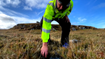 A person wearing a hi-vis jacket and a beanie hat is bending down in the foreground of the picture and grabbing at the peatland beneath his feet. Behind him is more peatland and a blue sky with wispy white clouds.