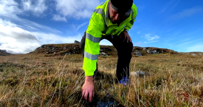 A person wearing a hi-vis jacket and a beanie hat is bending down in the foreground of the picture and grabbing at the peatland beneath his feet. Behind him is more peatland and a blue sky with wispy white clouds.