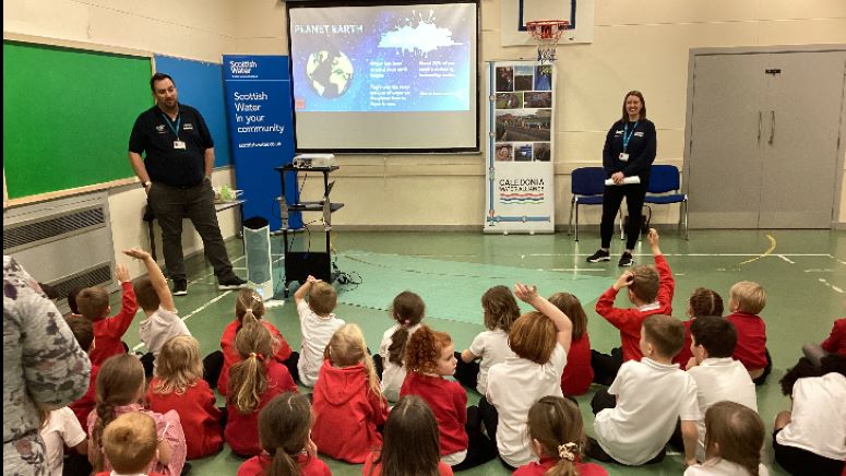 Two CWA employees stand in front of projector screen in front of a sat down group of pupils with hands up