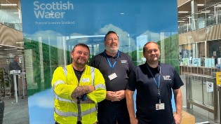 Field Response Clyde Team Celebrates Long-Service Three employees standing in front of a Scottish Water banner inside an office building. The person on the left is wearing a high-visibility vest, and the other two are in casual work attire, all smiling at the camera.
