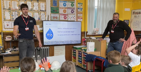 A man wearing Scottish Water uniform speaks in front of a class or primary pupils. Some of the pupils have their hands raised.