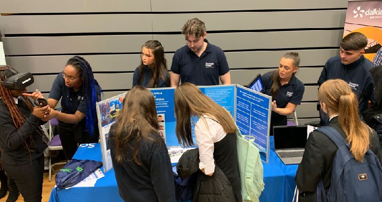 School pupils gather around a stall with a blue table cover and blue themed posters. Scottish Water staff stand behind talking and interacting with the pupils.