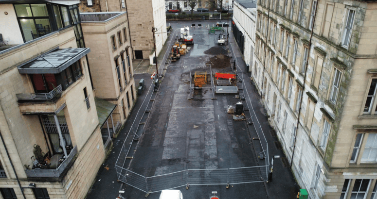 A construction site on a closed-off urban street between two rows of buildings. The street is fenced off with metal barriers, and construction equipment, including a digger, piles of dirt, and building materials, are visible on the asphalt surface. The buildings on either side are multi-story, made of light-colored stone, with some balconies and windows overlooking the site.
