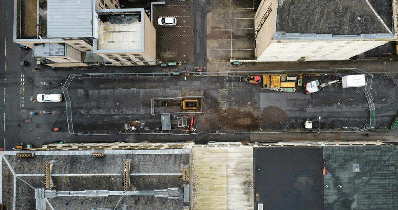 A construction site on a closed-off urban street between two rows of buildings. The street is fenced off with metal barriers, and construction equipment, including a digger, piles of dirt, and building materials, are visible on the asphalt surface. The buildings on either side are multi-story, made of light-colored stone, with some balconies and windows overlooking the site.