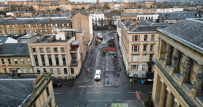 A construction site on a closed-off urban street between two rows of buildings. The street is fenced off with metal barriers, and construction equipment, including a digger, piles of dirt, and building materials, are visible on the asphalt surface. The buildings on either side are multi-story, made of light-colored stone, with some balconies and windows overlooking the site.