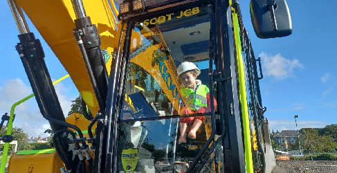 A child sits in a digger on a construction site. The sky in the background is blue.