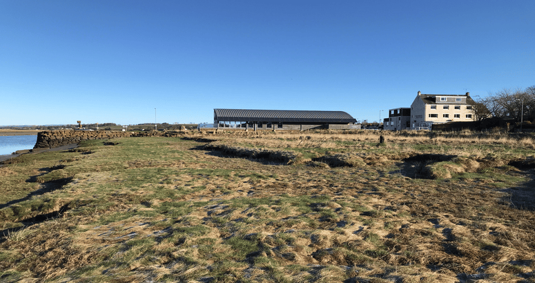 "A coastal landscape with grassy and marshy terrain in the foreground. A stone wall extends into the water on the left, and a modern boathouse with a slanted roof is visible in the middle ground. To the right, there is a large, multi-story white building with several windows. The sky is clear and blue, and the scene is illuminated by bright sunlight."