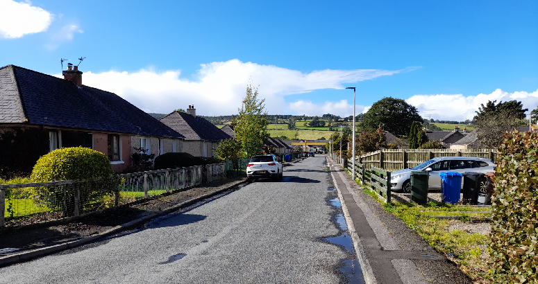 A quiet suburban street with single-story houses on either side, lined with fences and small gardens. A few cars are parked along the road. In the distance, green hills and trees stretch across the horizon under a bright blue sky with scattered clouds.