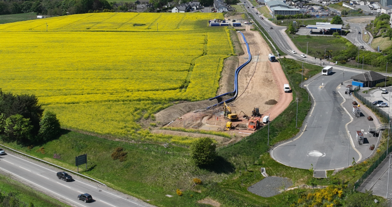 Aerial view of a vibrant landscape showing a large bright yellow rapeseed field alongside roads with moving vehicles and construction equipment near a developing area. Rolling hills and greenery are visible in the background under a clear sky.