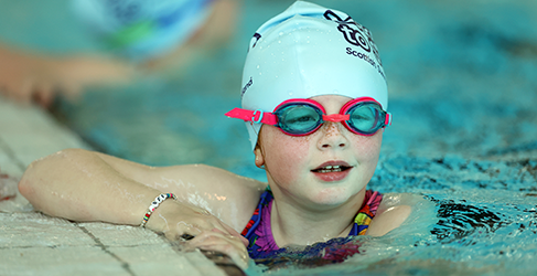 young girl with goggles and swim hat on in pool at poolside