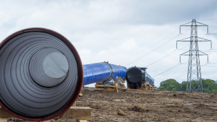 A ground-level photograph of an open water pipe upon the ground. The pipe is blue. In the background is an electricity pylon.