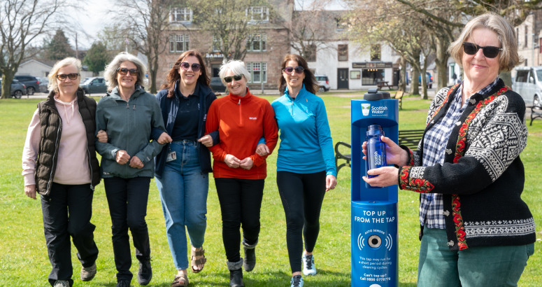 A group of people stand with their arms linked, facing towards the camera while standing on grassed area. On the right hand side, a person stands holding a water bottle and fills it under a blue Scottish Water top up tap.