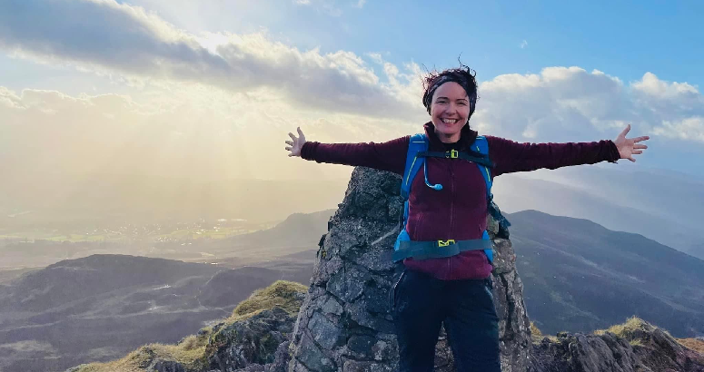 Jo Hedley celebrates reaching the top of a Munro. She smiles with her arms outstretched at the peak with blue skies and the sun shining behind her.