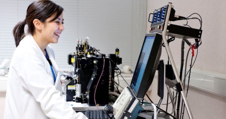 Scientist in lab coat using a laptop with advanced laboratory equipment in the background.
