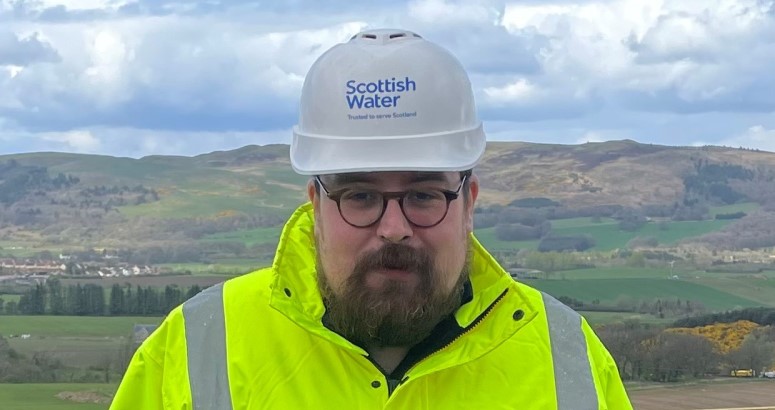 Person with beard posing with branded Scottish Water hard hat and hi-vis PPE, with lots of green hills in the background