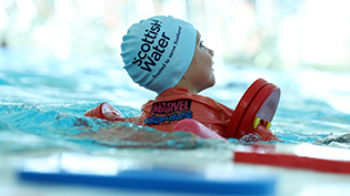 back view of young child in swimming pool with red float, wearing Learn to Swim cap