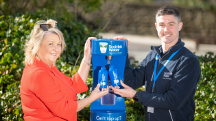 Two people stand at a blue Scottish Water top-up tap holding blue metal bottles branded in the Scottish Water logo.